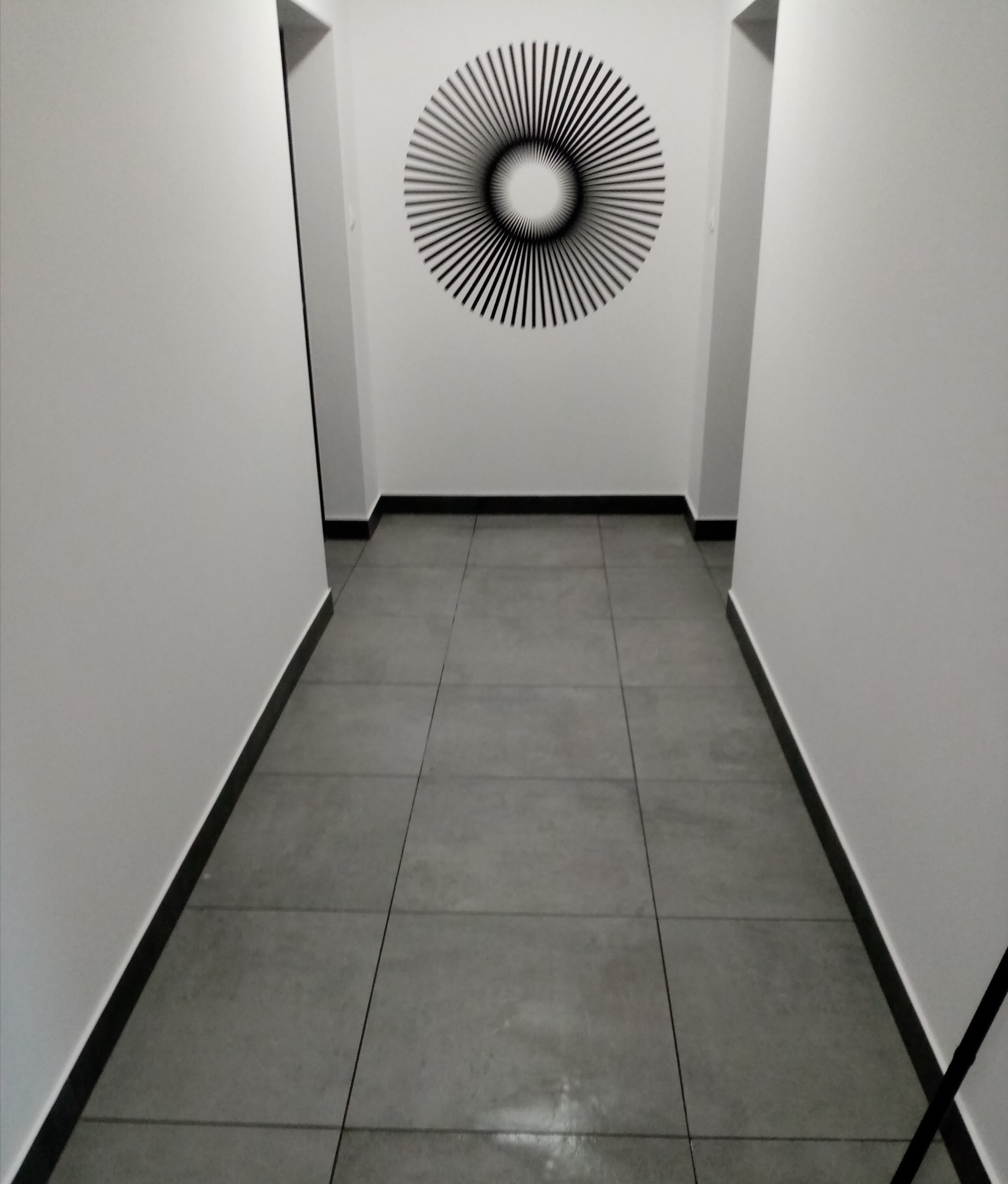 Wnętrze korytarza z szarymi płytkami podłogowymi, białymi ścianami i czarnymi listwami przypodłogowymi; na ścianie okrągła, abstrakcyjna dekoracja.