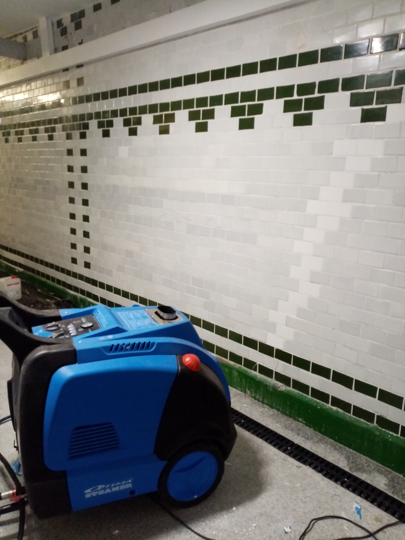 Niebieski parowy odkurzacz przemysłowy stoi na posadzce przed ścianą wyłożoną białymi kafelkami z zielonymi akcentami, widoczna kratka odpływowa w podłodze.