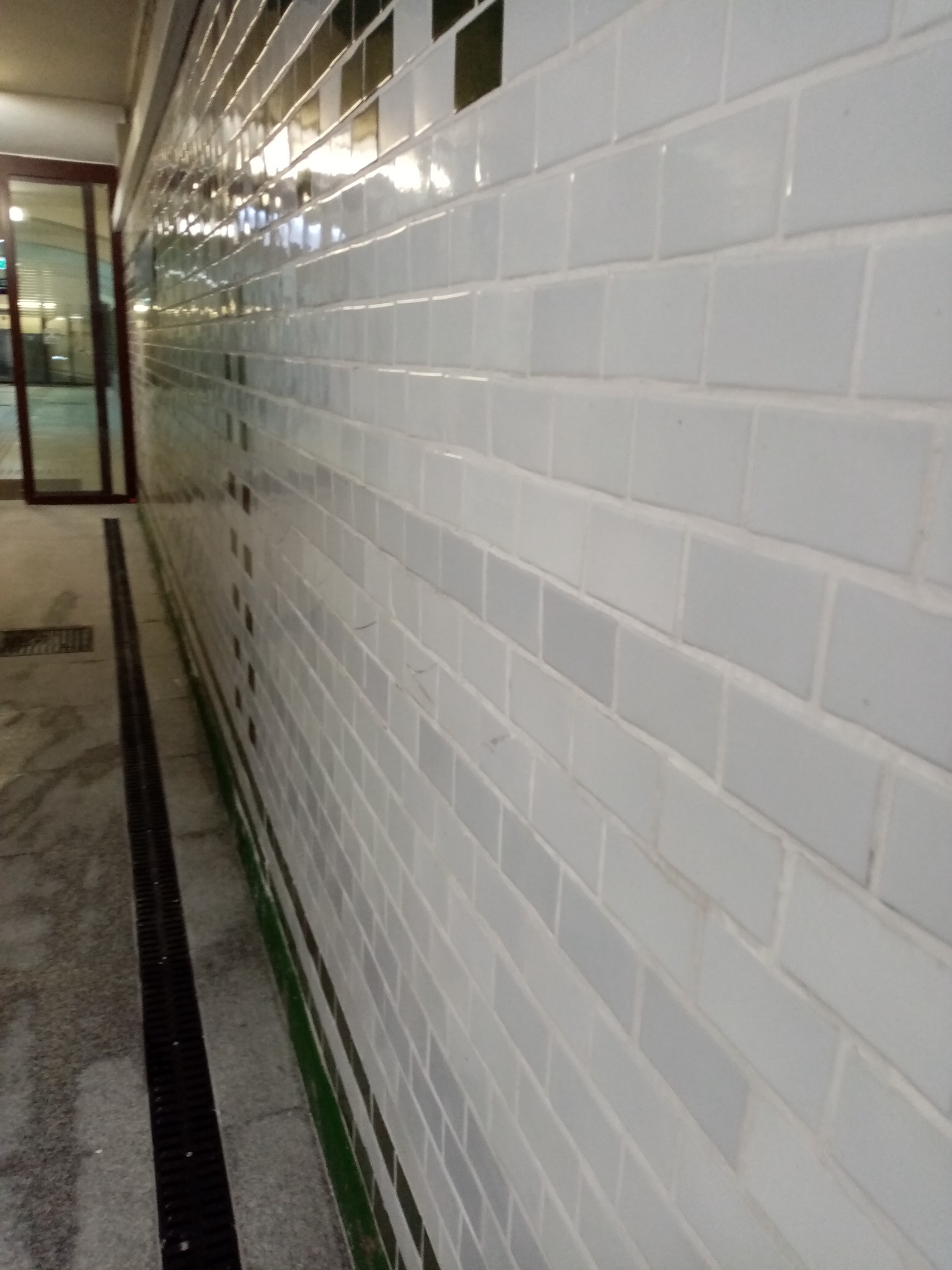 Białe, błyszczące kafelki na ścianie w przejściu, widoczna kratka odpływowa na podłodze i fragment drzwi na końcu korytarza.