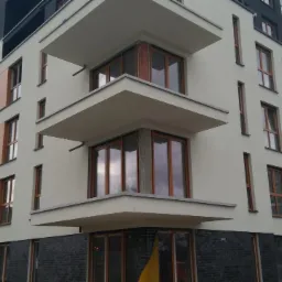 Nowoczesny budynek mieszkalny z jasną elewacją, drewnianymi oknami i balkonami. Dolna część wykończona ciemną cegłą. Widoczne detale architektoniczne.