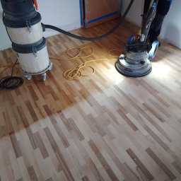 Posadzkarstwo Piotr Dąbrowa - Parkiet poddawany renowacji za pomocą cykliniarki przemysłowej; widoczne różnice w odcieniu drewna po procesie szlifowania, obok stoi odkurzacz przemysłowy.