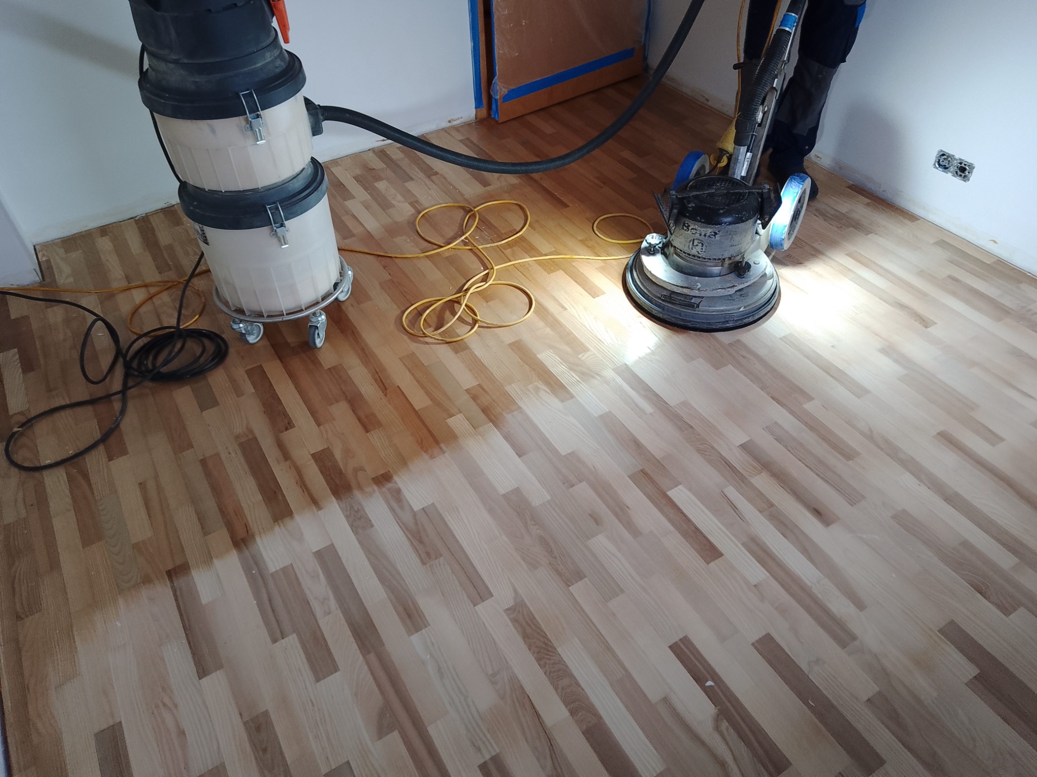 Parkiet poddawany renowacji za pomocą cykliniarki przemysłowej; widoczne różnice w odcieniu drewna po procesie szlifowania, obok stoi odkurzacz przemysłowy.