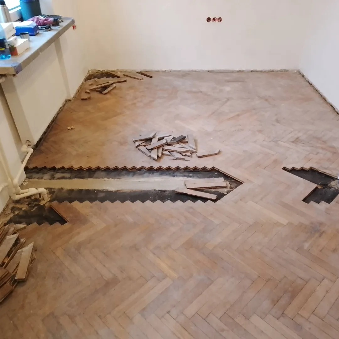 Demontaż parkietu ułożonego w jodełkę, widoczne fragmenty usuniętej podłogi i przygotowane deszczułki.
