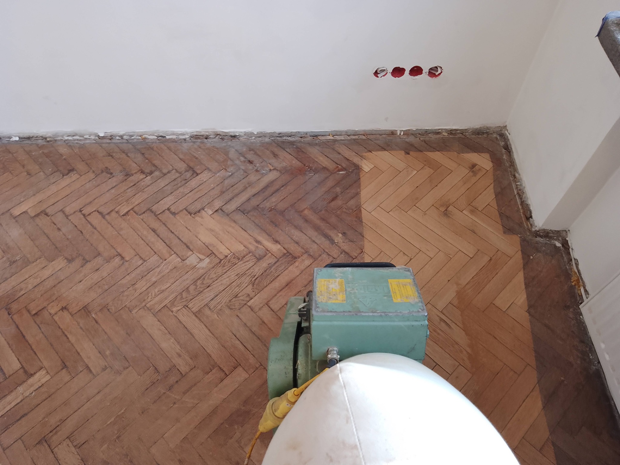 Cyklinowanie parkietu ułożonego w jodełkę; widoczna maszyna do cyklinowania i fragment podłogi przed i po cyklinowaniu.