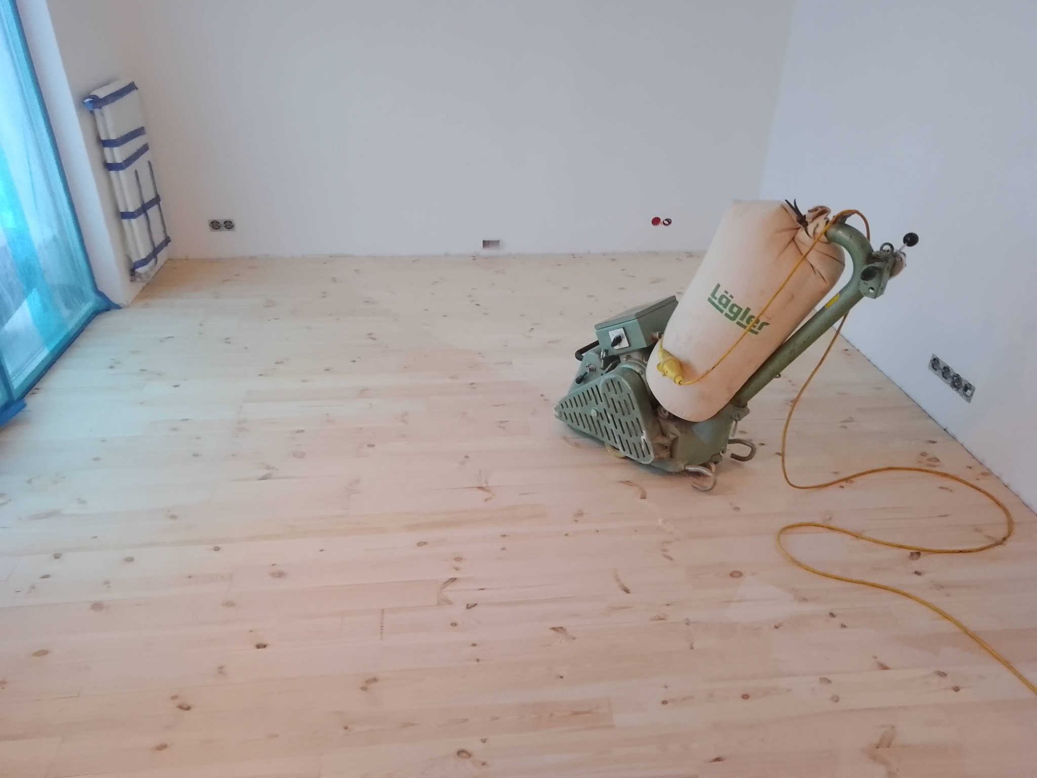 Parkiet z jasnego drewna po obróbce, widoczna szlifierka marki Lägler z workiem na pył, ściany zabezpieczone folią malarską.