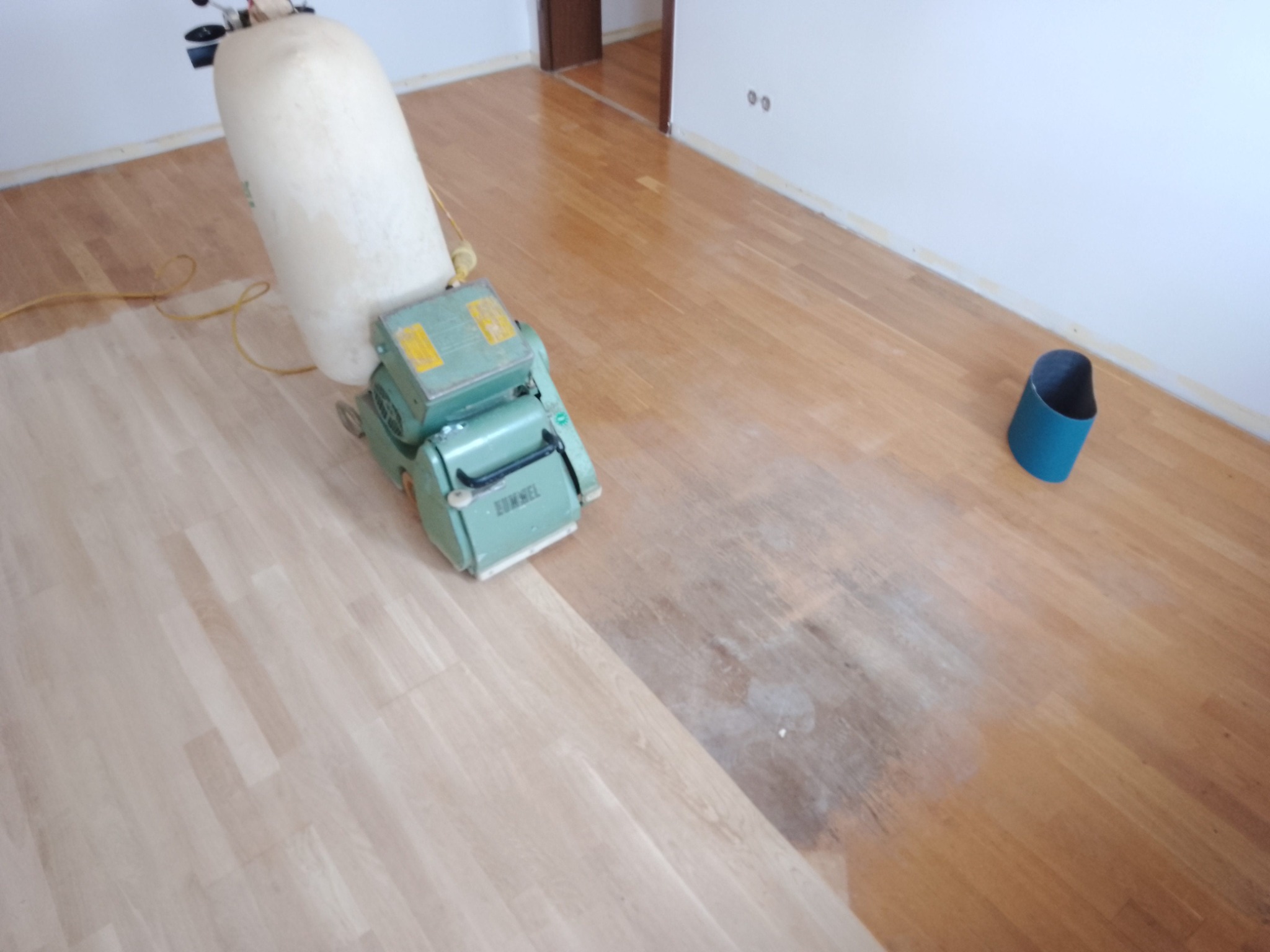 Maszyna do renowacji parkietu w trakcie pracy, widoczna różnica w kolorze między odnowioną a nieodnowioną częścią podłogi, obok ściany stoi niebieski pojemnik.
