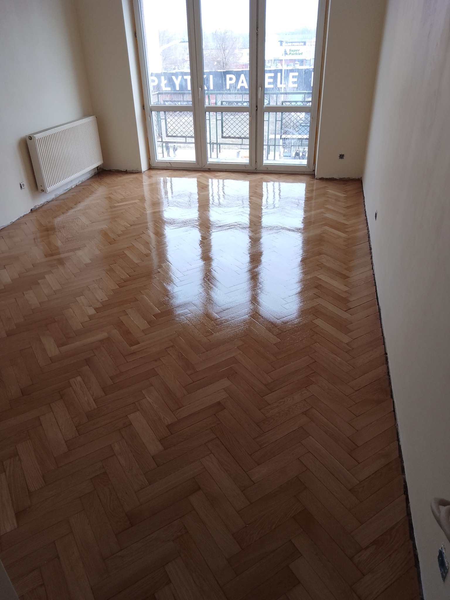 Świeżo polakierowany parkiet ułożony w jodełkę, z widocznym odbiciem okna balkonowego i kaloryfera, w jasnym pokoju po remoncie.