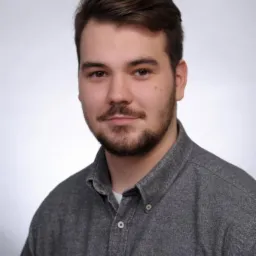 Portret młodego mężczyzny w szarej koszuli z brodą na jasnoszarym tle, z lekkim uśmiechem.