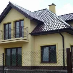Żółty dom dwukondygnacyjny z brązowym dachem i balkonem, otoczony ogrodzeniem z siatki. Widoczny komin i brama wjazdowa. Dom po odbiorze technicznym.