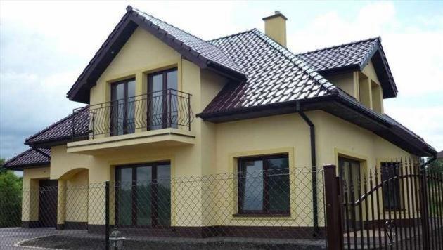 Żółty dom dwukondygnacyjny z brązowym dachem i balkonem, otoczony ogrodzeniem z siatki. Widoczny komin i brama wjazdowa. Dom po odbiorze technicznym.