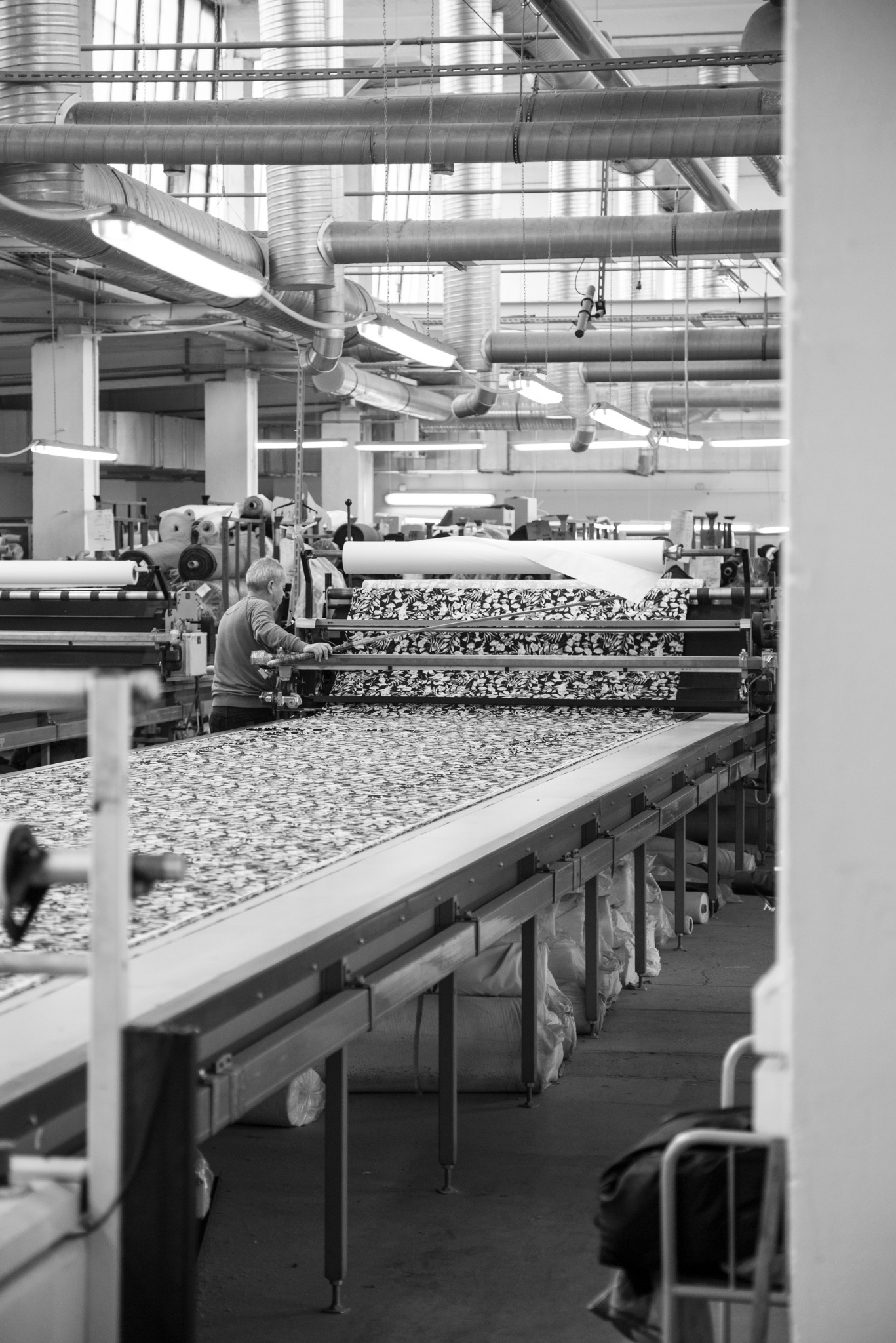 Czarno-białe zdjęcie szwalni z pracownikiem obsługującym maszynę do cięcia wzorzystych tkanin. Widoczne rury wentylacyjne pod sufitem i rolki materiałów w tle.
