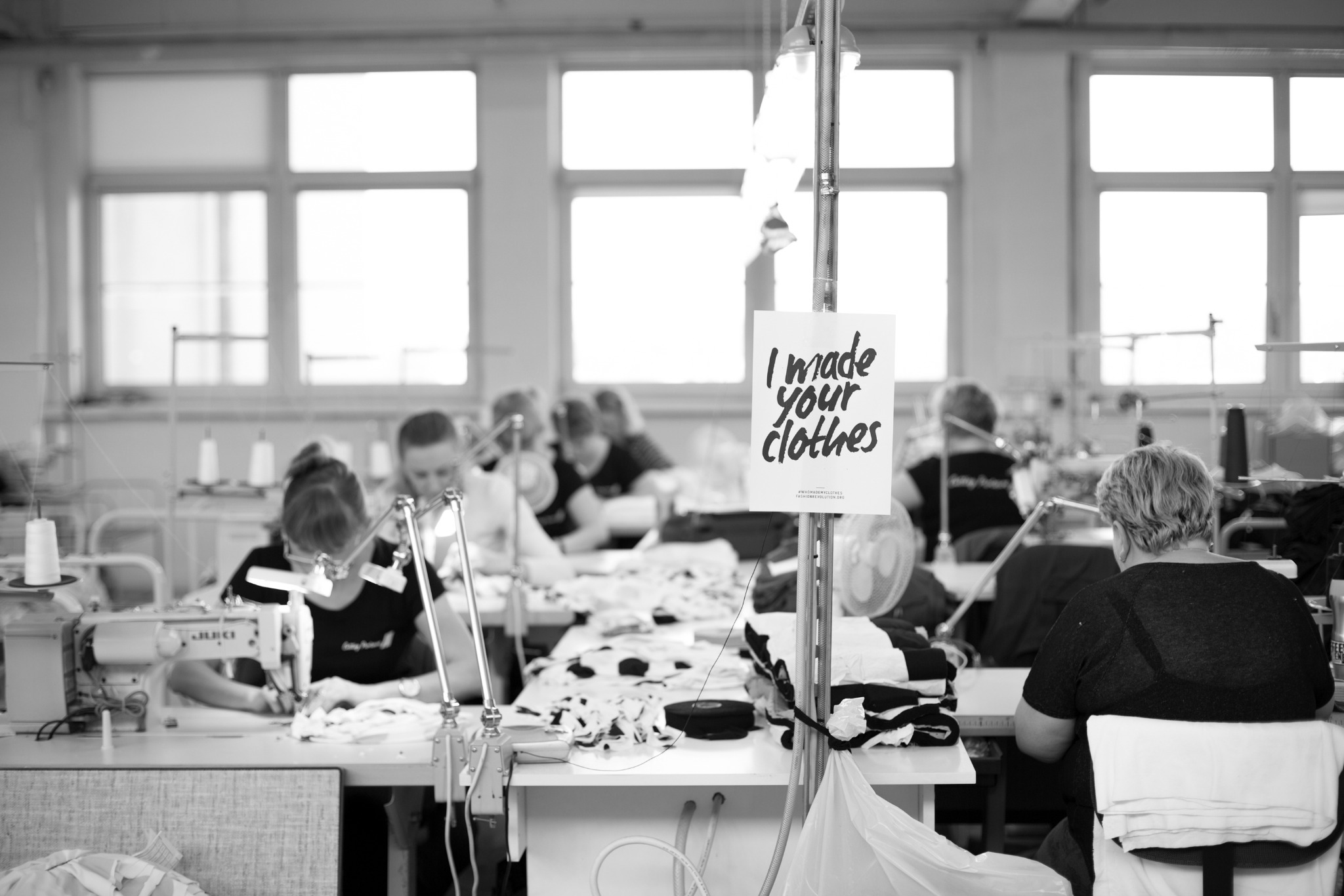 Czarno-białe zdjęcie szwalni: pracownicy szyją odzież przy maszynach, w tle okna i plakat 'I made your clothes'.