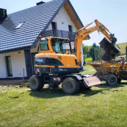 Żółta koparka Hyundai 55W-9A z łyżką nabiera ziemię i wysypuje do wywrotki na tle nowego domu z ciemnym dachem i zielonego trawnika.