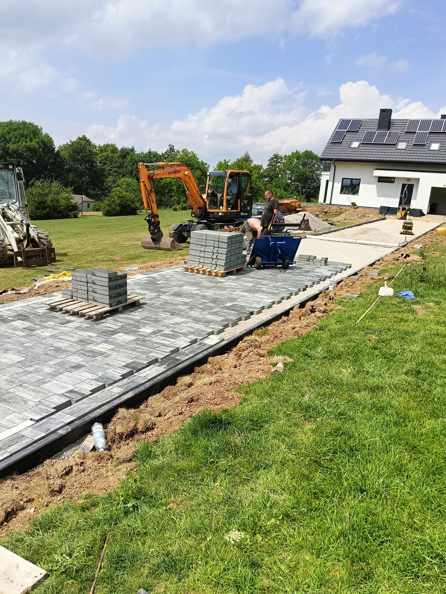 Montaż szarej kostki brukowej na podsypce piaskowej, z widocznym pracownikiem, wózkiem i mini koparką na tle domu z panelami słonecznymi.
