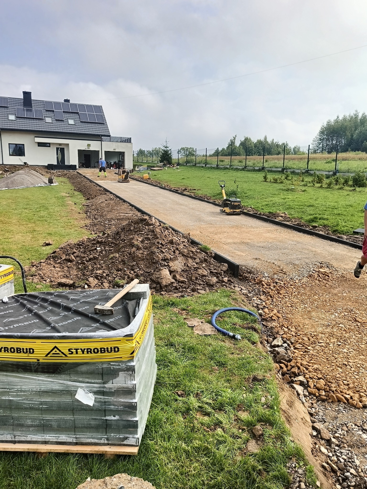 Przygotowanie podłoża pod nawierzchnię z kostki brukowej: ubijanie ziemi zagęszczarką, widoczne obrzeża, paleta z kostką Styrobud i młotek na trawie.