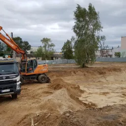 Pomarańczowa koparka Doosan nabiera ziemię do białej ciężarówki Volvo na placu budowy z widokiem na budynki mieszkalne w tle, oświetlenie pochmurne.