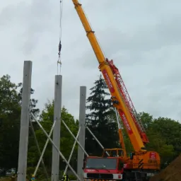 Żuraw wieżowy podnosi prefabrykowany słup betonowy na placu budowy z widocznymi fundamentami i pracownikami w kamizelkach odblaskowych.