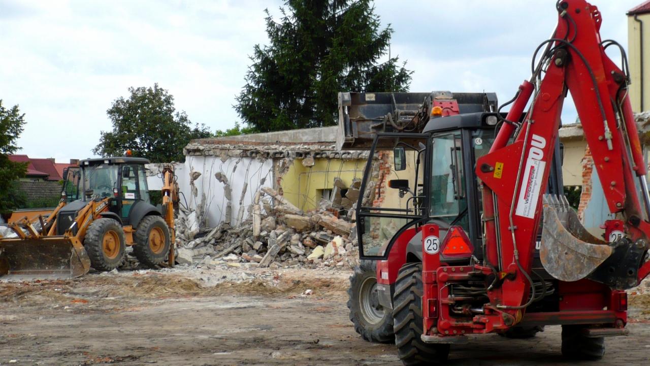 Dwa traktory z łyżkami na placu budowy, w tle zrujnowany budynek z fragmentami ścian i stertą gruzu.