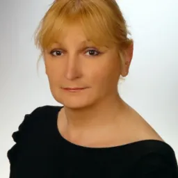 Portret kobiety o blond włosach, ubranej w czarną bluzkę na jasnym tle. Uśmiech na twarzy, bezpośrednie spojrzenie.