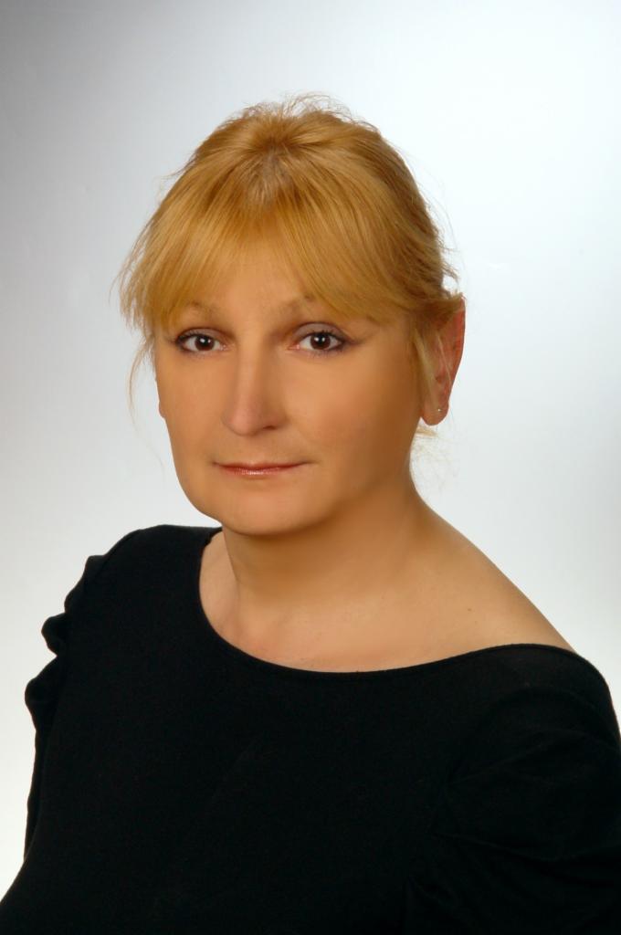 Portret kobiety o blond włosach, ubranej w czarną bluzkę na jasnym tle. Uśmiech na twarzy, bezpośrednie spojrzenie.