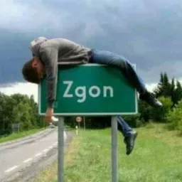 Mężczyzna w szarym ubraniu leży wyczerpany na zielonym znaku drogowym z napisem 'Zgon' na tle pochmurnego nieba i pobocza drogi.