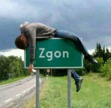 Mężczyzna w szarym ubraniu leży wyczerpany na zielonym znaku drogowym z napisem 'Zgon' na tle pochmurnego nieba i pobocza drogi.