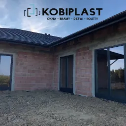 Surowe ściany nowego domu z zamontowanymi oknami w ciemnych ramach, widoczne logo firmy Kobiplast, dach pokryty ciemną dachówką.