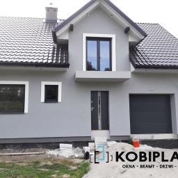 Kobiplast S.C - Nowo wybudowany dom jednorodzinny z szarą elewacją, czarną dachówką i czarnymi oknami. W tle widoczny fragment ogrodu. Przed domem stos materiałów budowlanych.