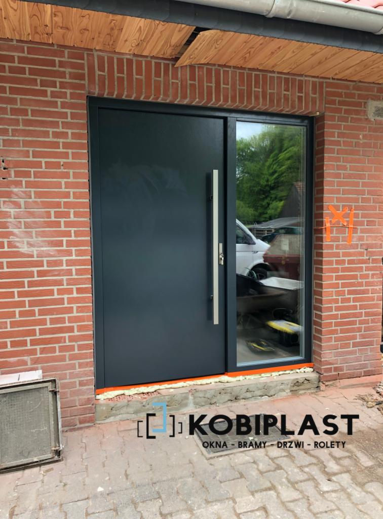 Nowoczesne, antracytowe drzwi wejściowe z długim, pionowym uchwytem i szybą boczną, zamontowane w murze z czerwonej cegły, widoczne ślady montażu i logo firmy Kobiplast.