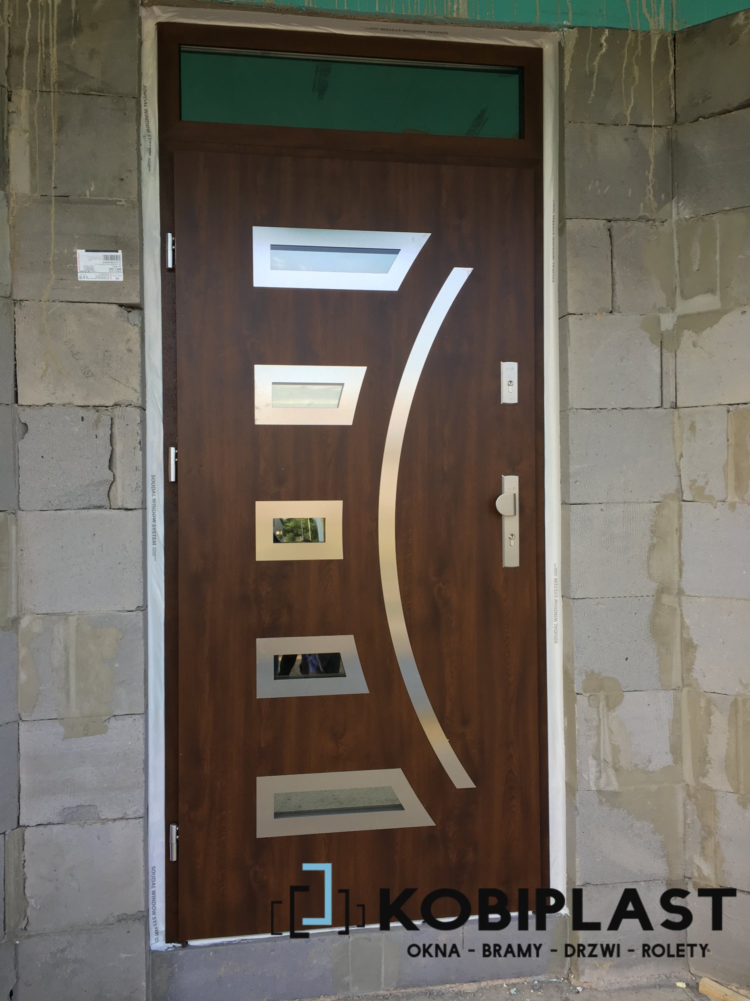 Nowoczesne drzwi wejściowe z przeszkleniami i srebrnym elementem dekoracyjnym, zamontowane w surowym murze z betonu komórkowego, widoczne logo firmy Kobiplast.