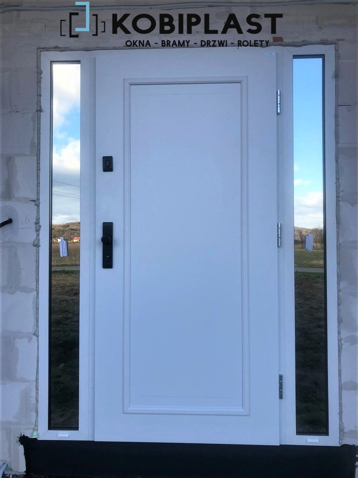Białe drzwi wejściowe z bocznymi oknami, wstawione w surowy mur z pustaków, widoczne logo firmy Kobiplast w górnej części.