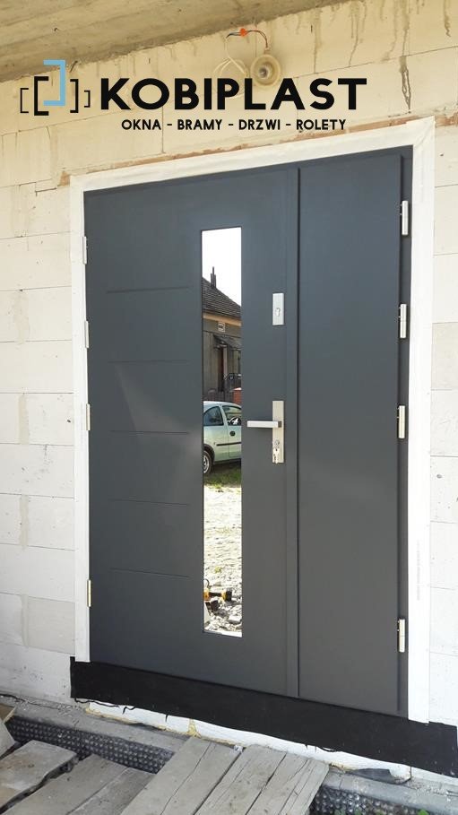 Szare drzwi wejściowe z szybą zamontowane w surowym stanie, widoczny budynek w tle przez szybę.