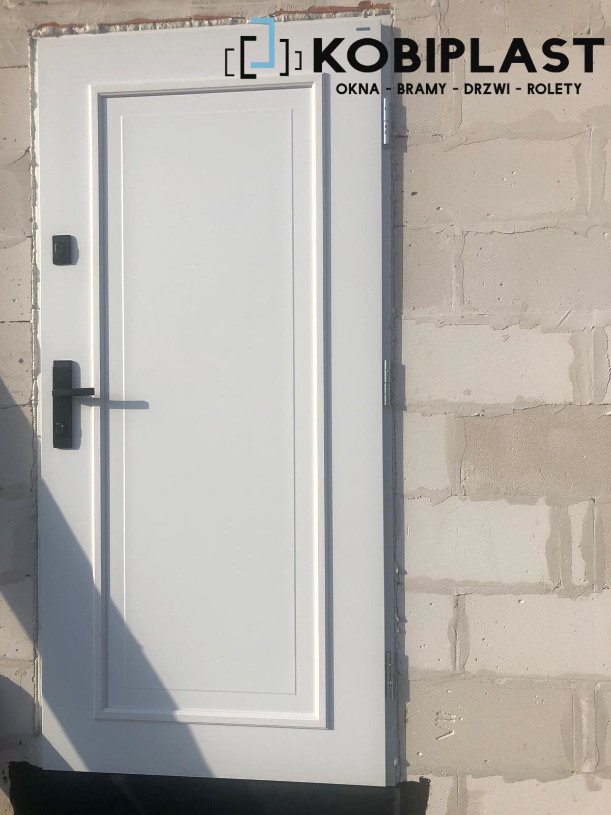 Białe drzwi wejściowe z czarną klamką i zamkiem, zamontowane w surowej ścianie z betonu komórkowego, z widocznym logo producenta u góry.