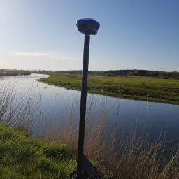 Urządzenie pomiarowe CHCNAV na statywie, ustawione na brzegu rzeki z trzcinami, w tle rozległe łąki i las pod błękitnym niebem.