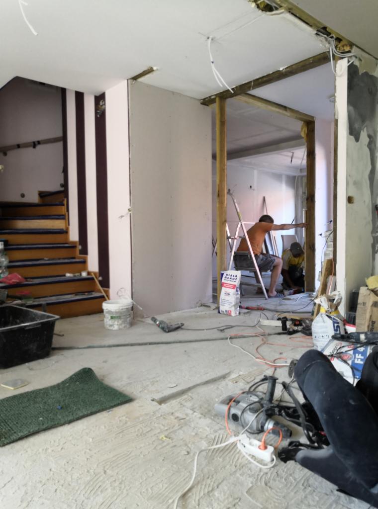 Wnętrze w trakcie remontu: montaż płyty gipsowo-kartonowej, widoczny pracownik na drabinie, narzędzia, worki z gładzią, nieporządek na podłodze, fragment schodów w tle.