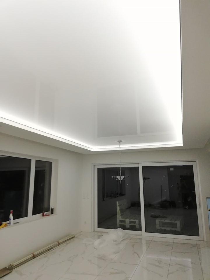 Biały, błyszczący sufit napinany z oświetleniem LED wzdłuż krawędzi, widok na marmurowe płytki podłogowe i przesuwne drzwi balkonowe.