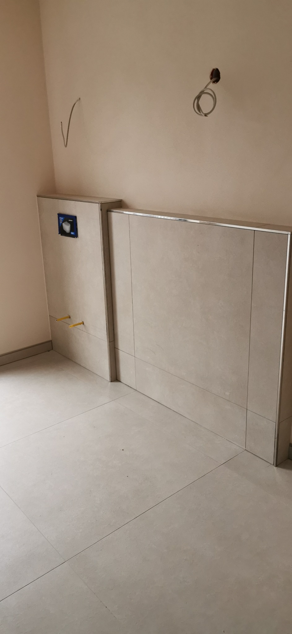 Narożne ujęcie fragmentu łazienki z zabudowanym stelażem podtynkowym WC, wykończonym beżowymi płytkami wielkoformatowymi z metalową listwą, widoczne wystające rurki oraz otwór na kabel elektryczny.