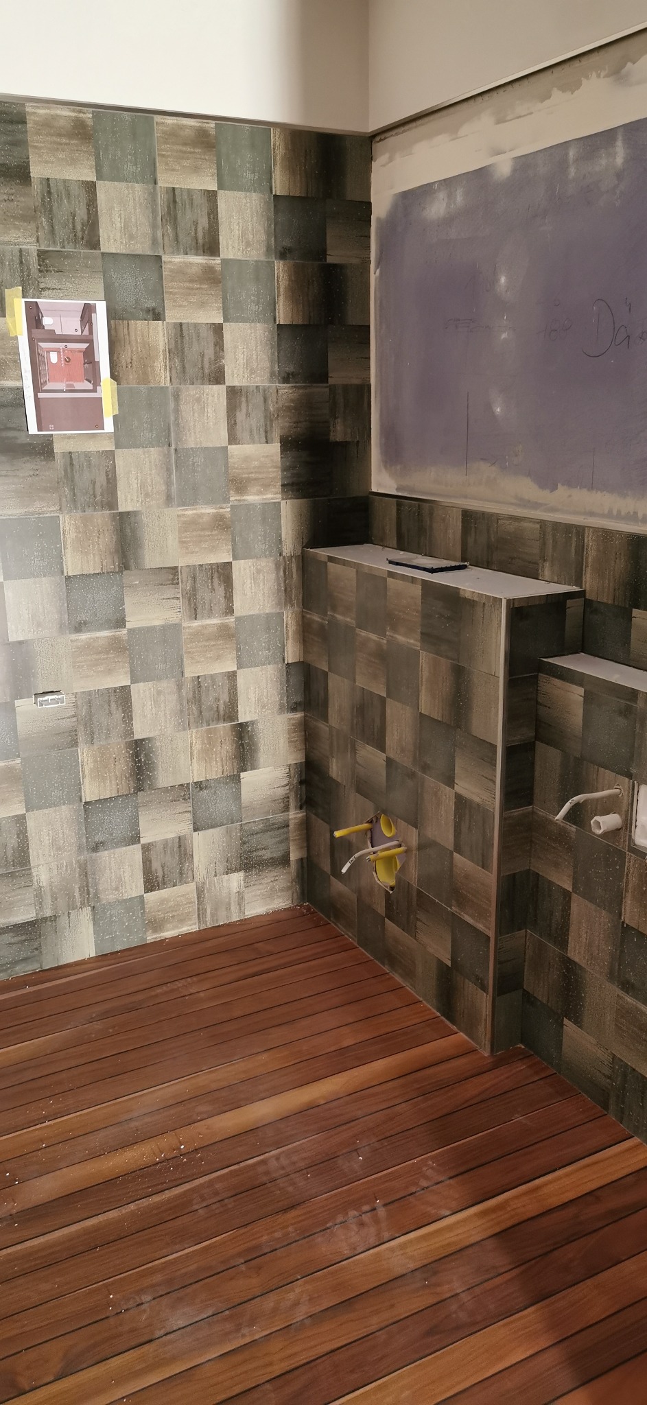 Wnętrze łazienki w trakcie prac wykończeniowych: ułożona podłoga z ciemnego drewna, ściany wyłożone mozaiką w odcieniach szarości i brązu, widoczne wystające rury instalacji wodnej, na ścianie...