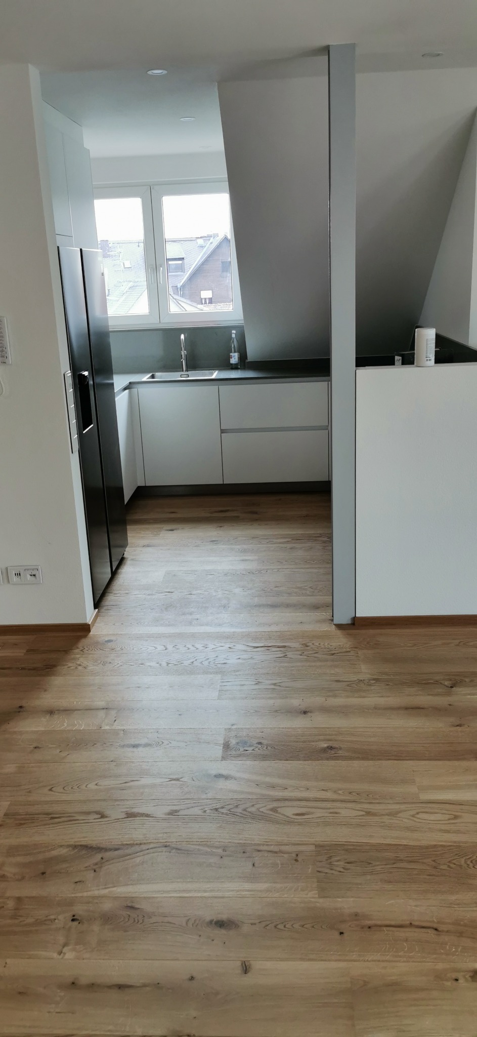 Nowoczesna, jasna kuchnia w mieszkaniu na poddaszu z drewnianą podłogą, białymi szafkami i czarną lodówką.