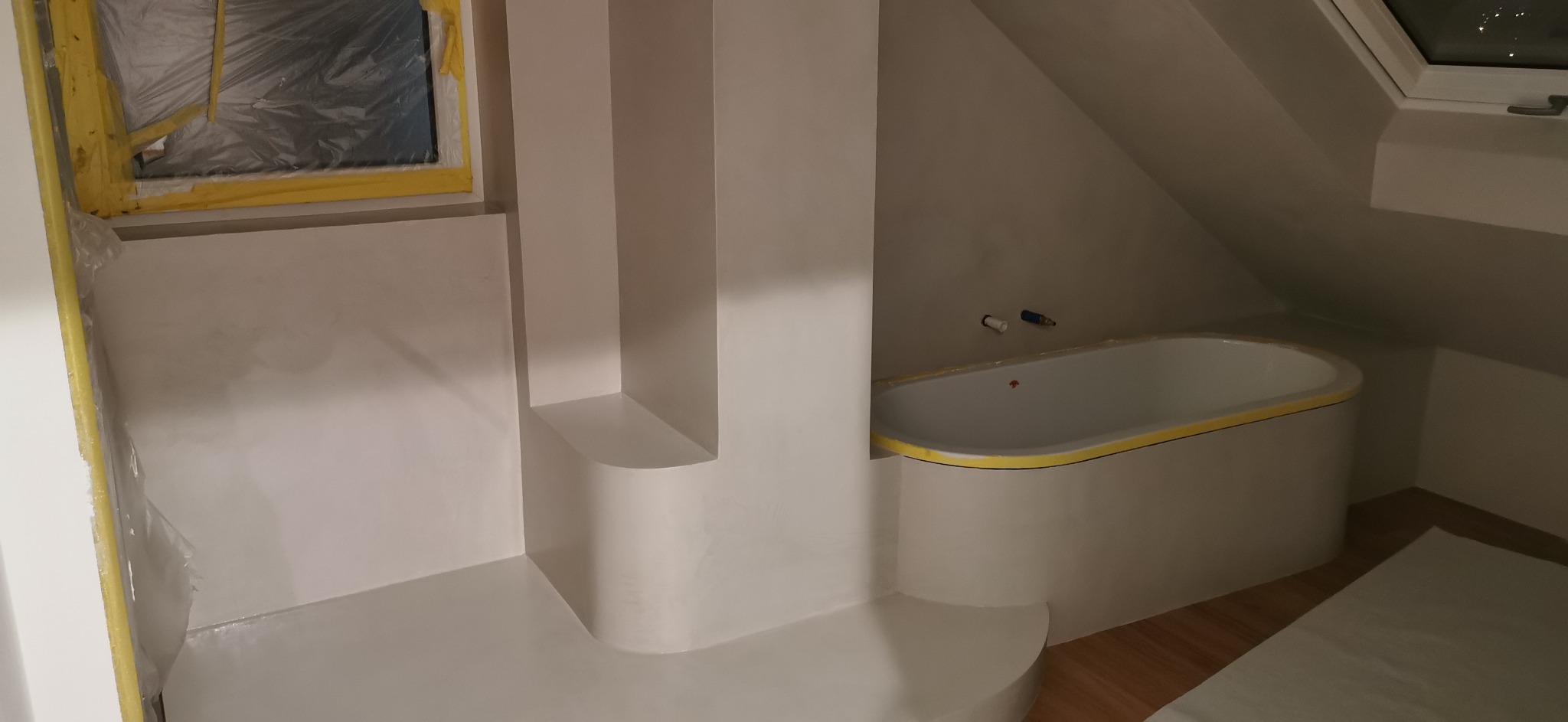 Łazienka na poddaszu w trakcie remontu: świeżo pomalowane ściany na biało, zabezpieczone okno folią i taśmą malarską, wanna obłożona taśmą, widoczne przyłącza wodne.