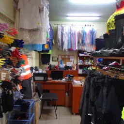 Wnętrze sklepu z odzieżą roboczą, prezentacja rękawic ochronnych na ścianie, obok wieszaki z ubraniami, na półkach buty robocze, w tle biurko z komputerem i manekinem.