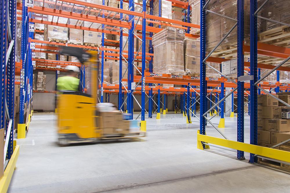 Wnętrze magazynu z wysokim składowaniem, wózek widłowy w ruchu transportuje kartony, widoczne regały z towarem