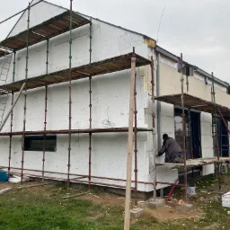 Budowa domu: robotnik na rusztowaniu montuje płyty izolacyjne z widoczną nakładką styropianową na elewacji, drugi robotnik obserwuje, w tle dom w stanie surowym zamkniętym.