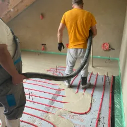 Wylewka samopoziomująca rozprowadzana na ogrzewaniu podłogowym przez dwóch pracowników w roboczych ubraniach i gumowcach, widoczne czerwone rury grzewcze i metalowe wsporniki.
