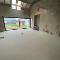 Wnętrze surowego domu jednorodzinnego z dużym oknem, widoczne ściany po tynkowaniu, wylewka na podłodze, drabina oparta o ścianę i elementy instalacji elektrycznej wystające ze ścian.