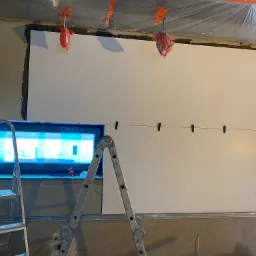 Montaż wielkoformatowych płyt na ścianie, widoczne poziomowanie za pomocą sznurka i klamerek, drabina aluminiowa oparta o ścianę, przygotowane otwory na gniazdka elektryczne.