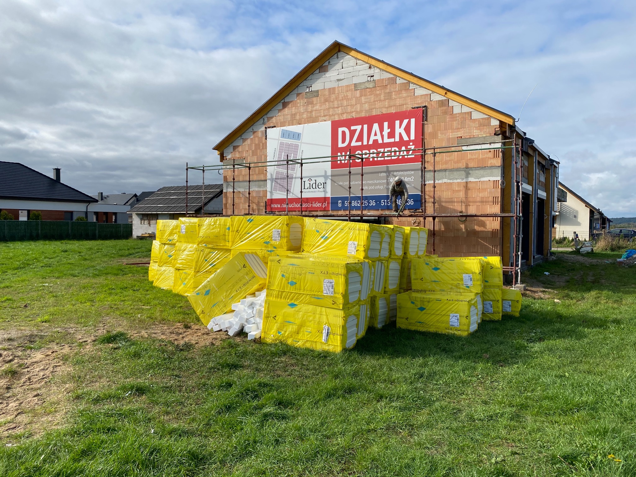 Budynek w trakcie budowy w Lęborku, z widocznymi rusztowaniami i stertą żółtych płyt termoizolacyjnych PIR na trawie. Na elewacji baner reklamowy z napisem 'Działki na sprzedaż'.
