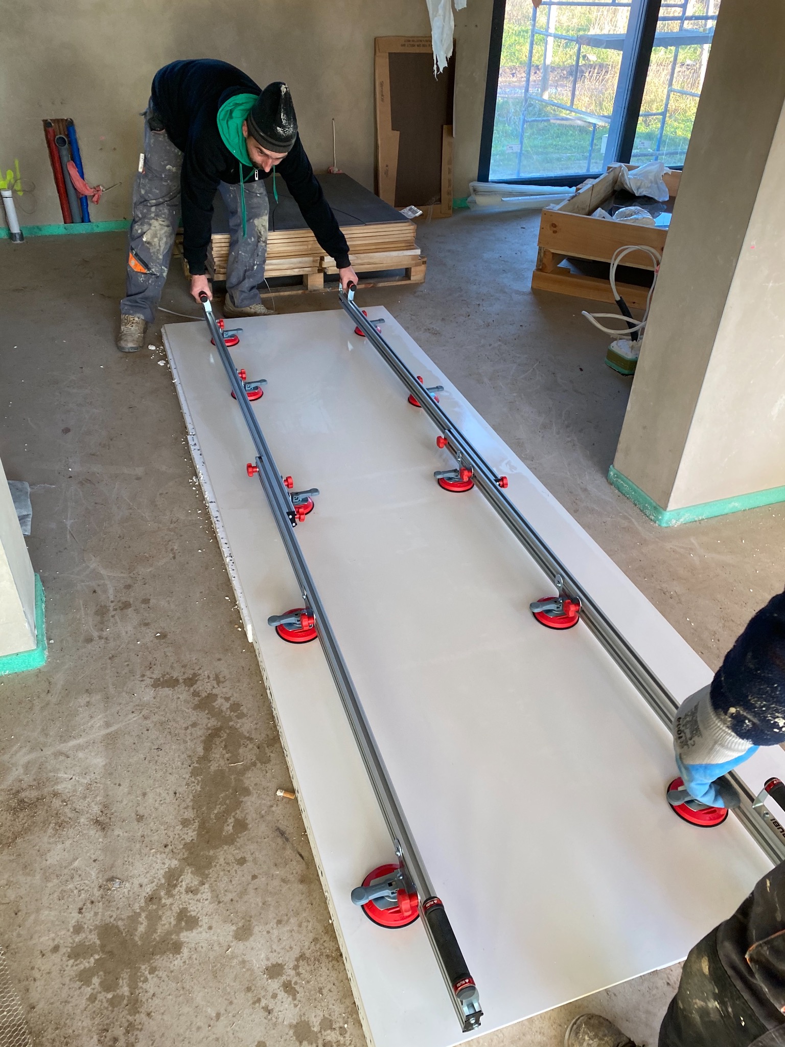 Dwóch mężczyzn przenosi dużą, białą płytę za pomocą specjalistycznych przyssawek i szyn prowadzących w surowym wnętrzu budynku w trakcie prac wykończeniowych.