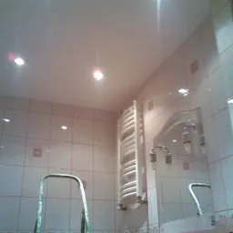 Wnętrze łazienki z jasnymi płytkami i dekoracyjnymi akcentami, widoczny grzejnik drabinkowy, fragment drabiny malarskiej i lustro z oświetleniem punktowym.