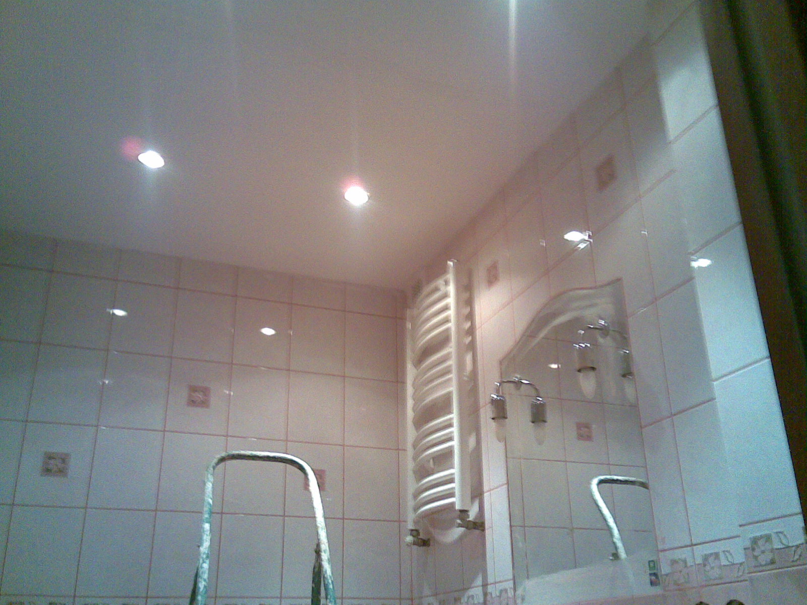 Wnętrze łazienki z jasnymi płytkami i dekoracyjnymi akcentami, widoczny grzejnik drabinkowy, fragment drabiny malarskiej i lustro z oświetleniem punktowym.